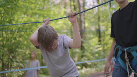 Children-on-a-summer-camp-hike-are-moving-along-the-ropes-with-the-help-of-a-guide-who-teaches-children-rock-climbing-and-tourism.-A-boy-in-the-forest-overcomes-a-rope-barrier
